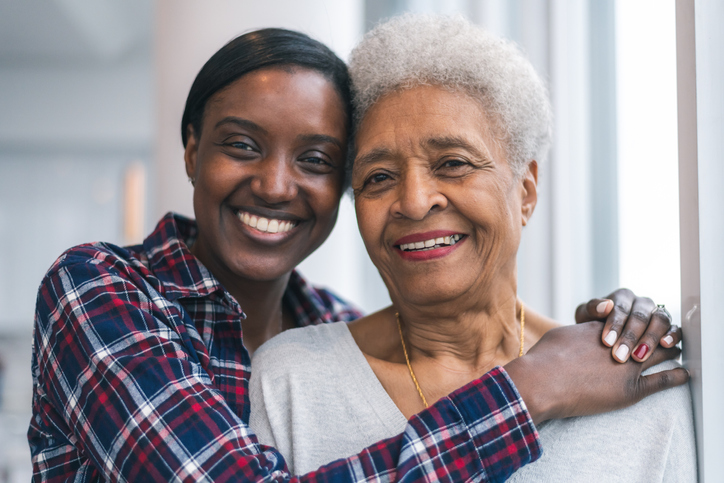 senior woman stands with her adult daughter, whose arms are wrapped around her shoulders, both smiling