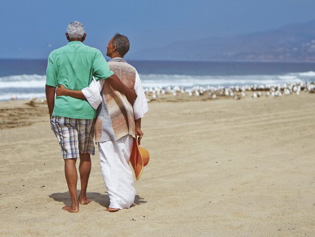 senior couple with their arms wrapped around one another look out at the ocean while enjoying a day at the beach