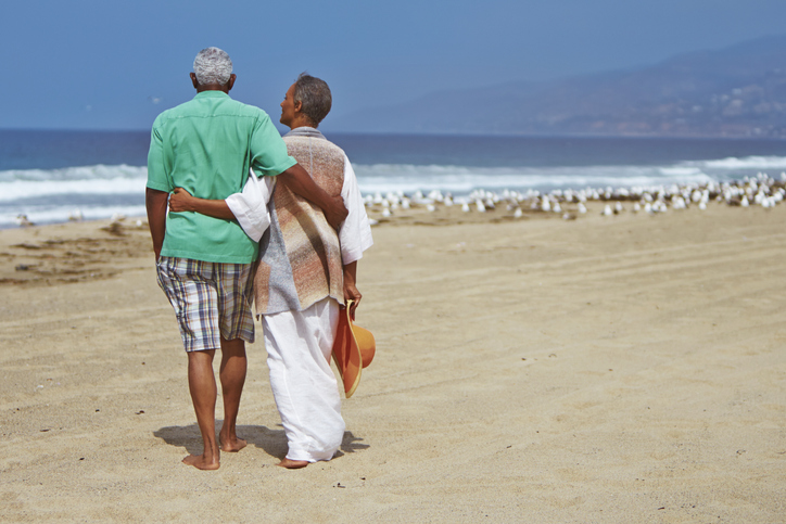 senior couple with their arms wrapped around one another look out at the ocean while enjoying a day at the beach