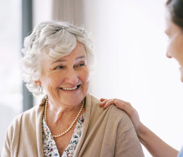 seated senior woman smiles at her female caregiver, who places her hand gently on her shoulder