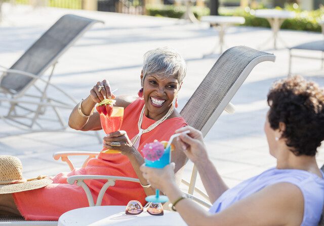 two senior women with umbrella adorned cocktails recline in poolside chairs, smiling