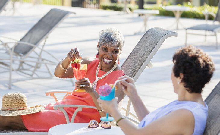 two senior women with umbrella adorned cocktails recline in poolside chairs, smiling