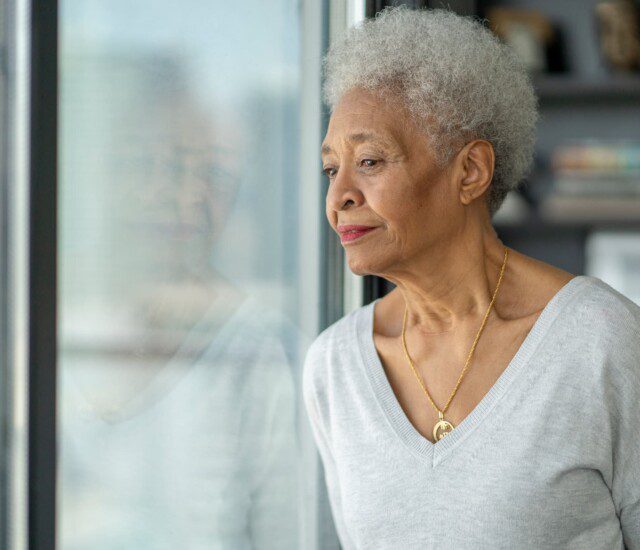 senior woman leans against a glass door in her home, looking out