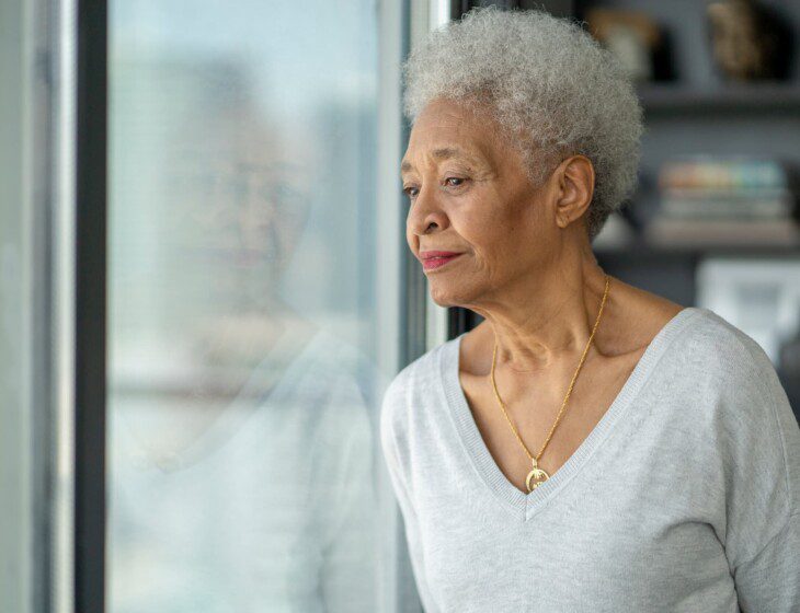 senior woman leans against a glass door in her home, looking out