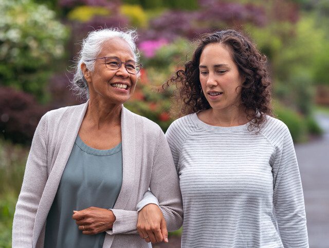 senior woman and her adult daughter smile and walk arm-in-arm together on a scenic path outdoors