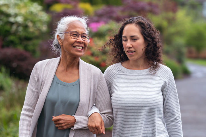 senior woman and her adult daughter smile and walk arm-in-arm together on a scenic path outdoors