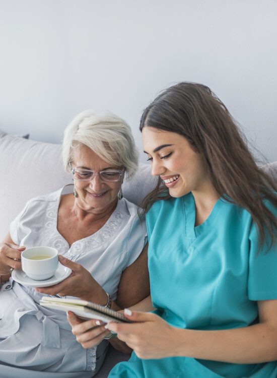 senior woman drinking coffee on her bed is shown information by her caretaker while seated