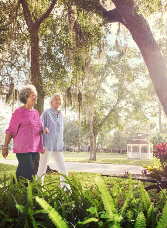two senior women walk together along a scenic path lined with palm trees and foliage at Village on the Green Senior Living Community