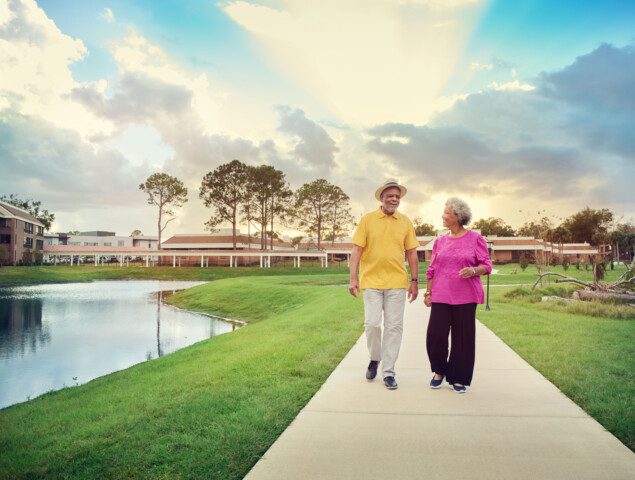 senior couple walks during sunset along a scenic path by the water at Village on the Green Senior Living Community