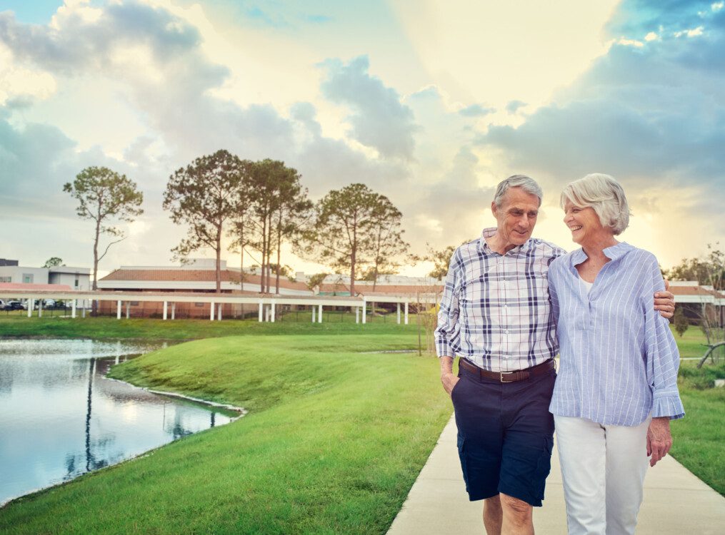 senior couple walks during sunset along a scenic path by the water at Village on the Green Senior Living Community