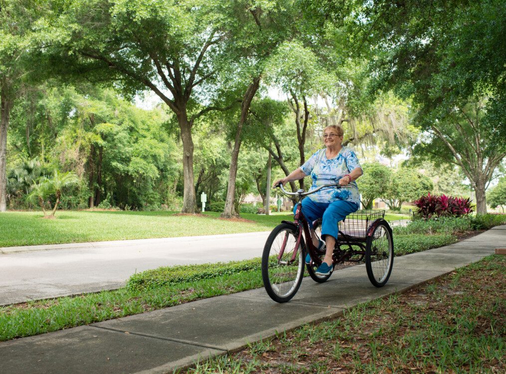 smiling senior woman in casual clothes rides a 3-wheel bike on a sidewalk path surrounded by trees