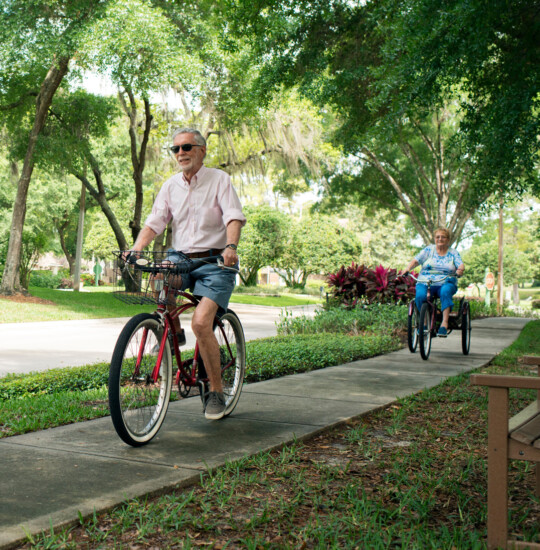 senior man with sunglasses rides bicycle on tree-lined sidewalk, with his wife close behind on a 3-wheel bike
