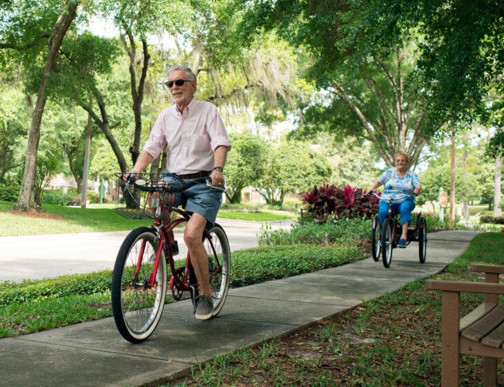 senior man with sunglasses rides bicycle on tree-lined sidewalk, with his wife close behind on a 3-wheel bike