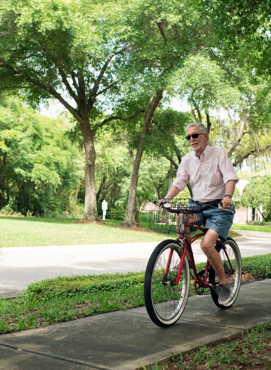 senior man with sunglasses rides bicycle on tree-lined sidewalk, with his wife close behind on a 3-wheel bike