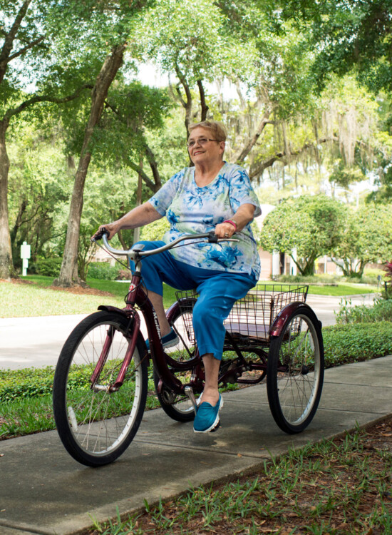 smiling senior woman in casual clothes rides a 3-wheel bike on a sidewalk path surrounded by trees