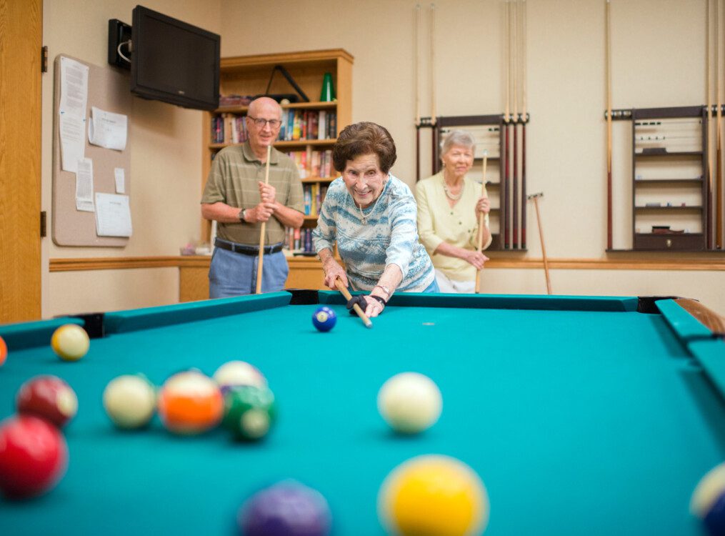 senior woman plays game of pool with her friends at their senior living community