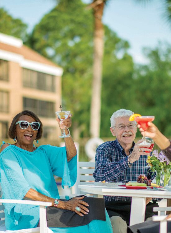 group of stylish and happy seniors toast cocktails together as they lounge by the pool at their senior living community