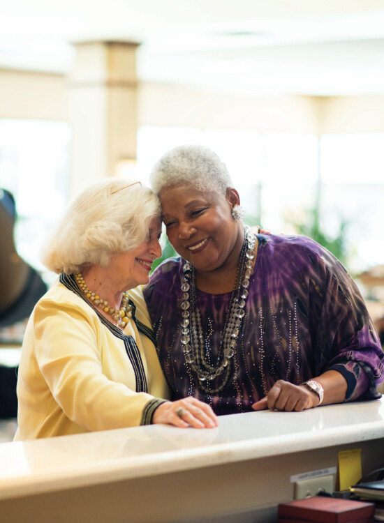 two senior woman smile from behind a tall counter, backdropped by a lounge