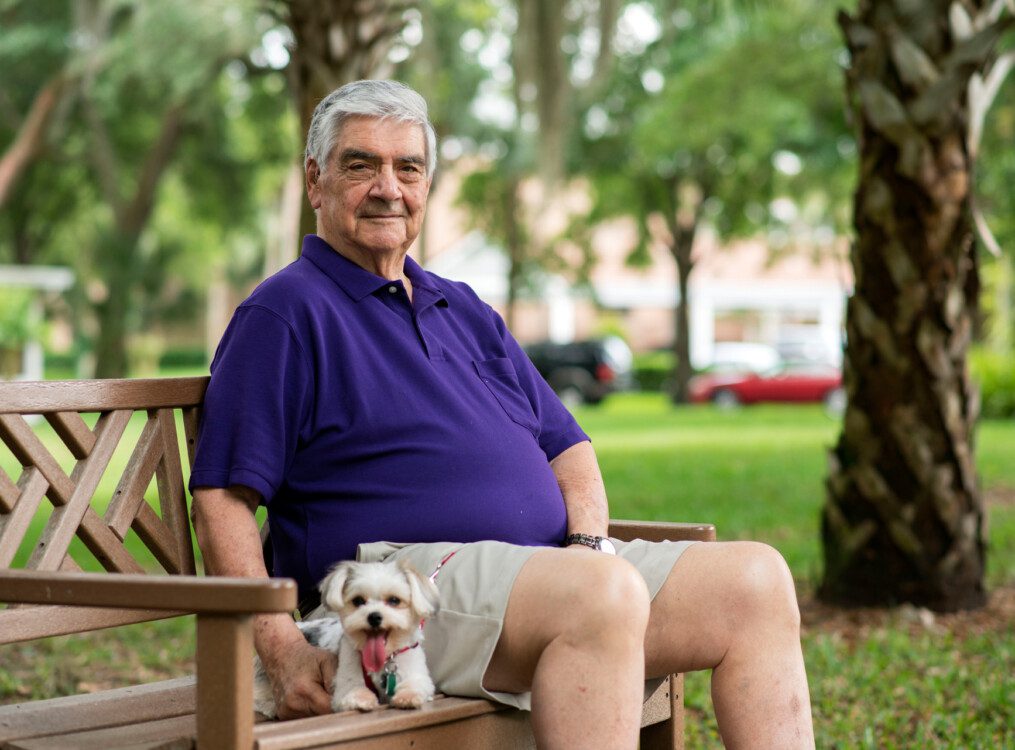 senior man sits with his small dog on a bench outside of his senior living community, backdropped by trees