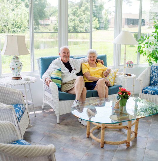 cute senior couple in casual clothes sit together on a loveseat in the patio of their villa, with large windows behind them