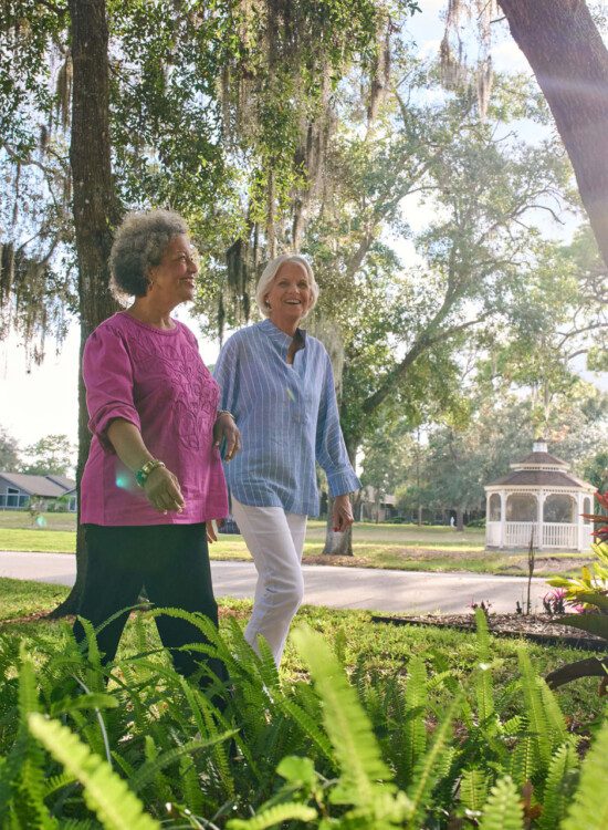 two senior women walk together along a scenic path lined with palm trees and foliage at Village on the Green Senior Living Community