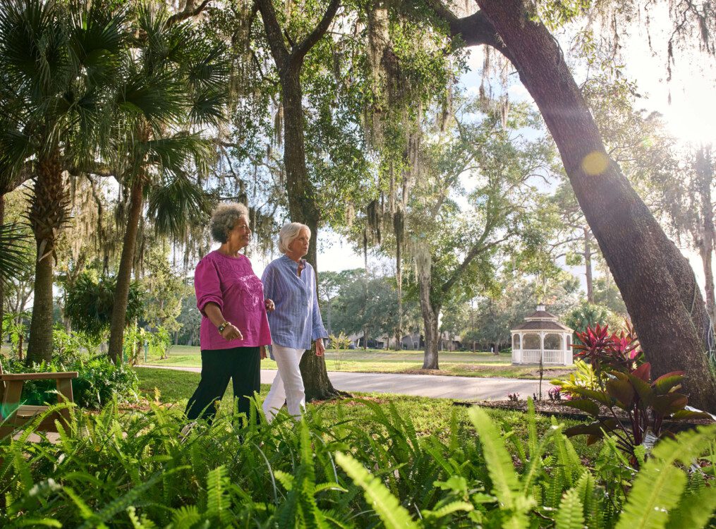 two senior women walk together along a scenic path lined with palm trees and foliage at Village on the Green Senior Living Community