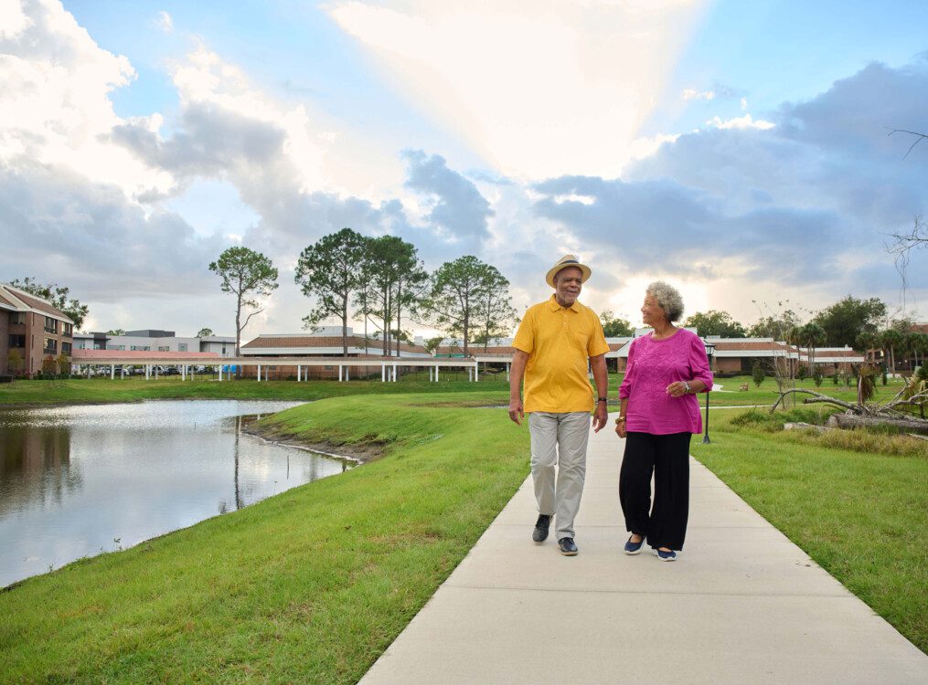 senior couple walks during sunset along a scenic path by the water at Village on the Green Senior Living Community