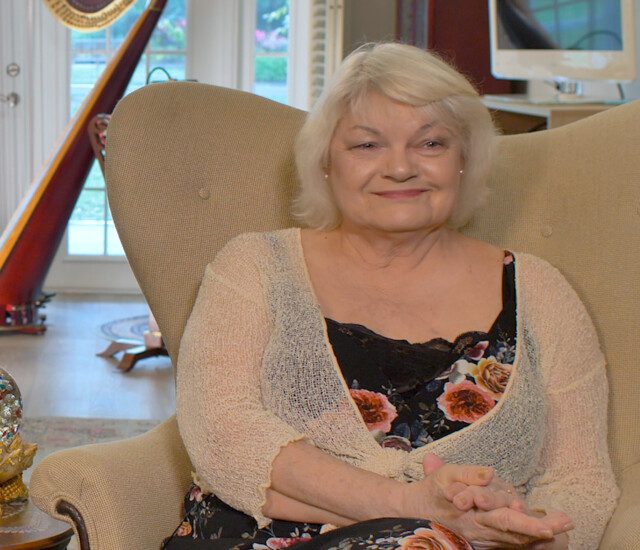 senior woman (Elizabeth Seabrooks) sits smiling in a chair, a harp behind her